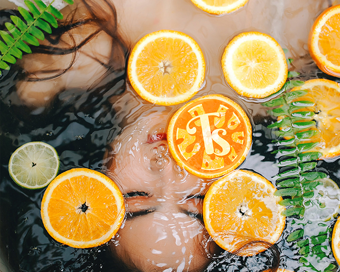Tropical splash logo mark on top of a photo featuring a girl sitting with her head underwater. Slices of oranges are floating on the water.