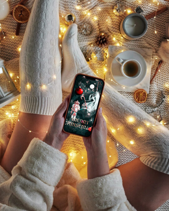 Christmas card mockup depicting a Christmas love story presented on a mobile screen, hold in hands by a young woman wearing long winter socks. Next her legs there are Christmas tree lights, pine cones, orange slices, vanilla sticks and a cup of coffee. Cosy Christmas atmosphere