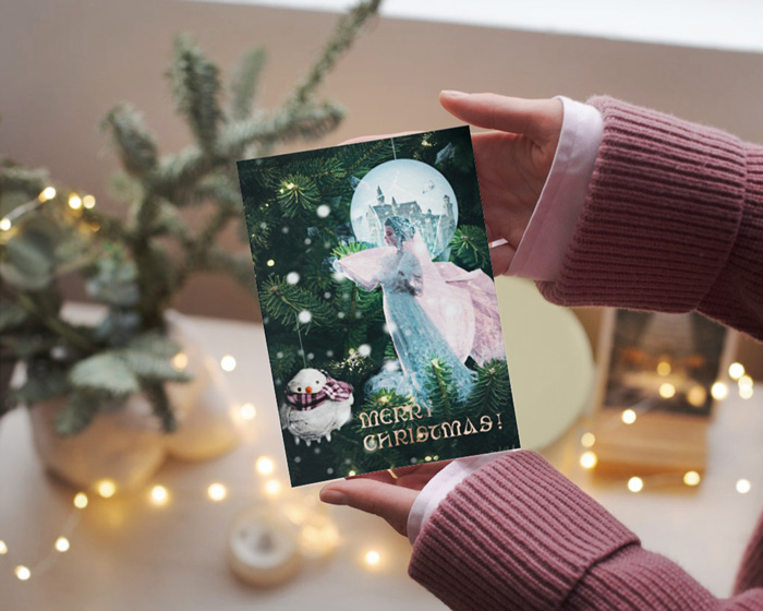 Christmas card mockup depicting The frozen kingdom presented on a Christmas card, held in hands by a woman. Behind there are Christmas tree branches and Christmas tree lights