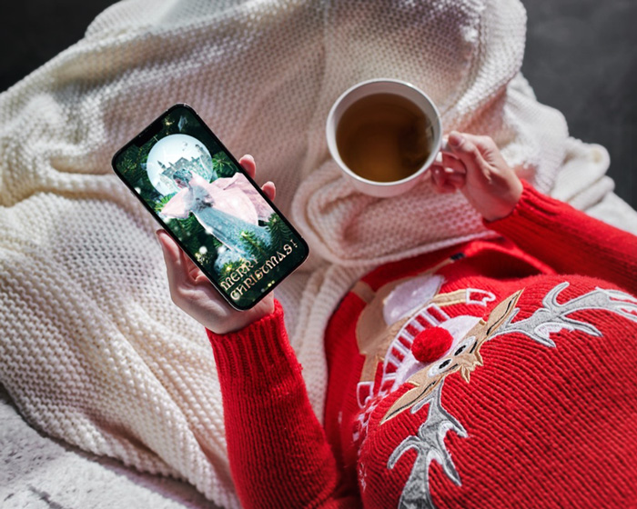 Christmas card mockup depicting The frozen kingdom presented on a mobile screen, held in hands by a woman wearing a sweater with a reindeer. The woman holds in her hand a cup with tea and her legs are covered with a blanket