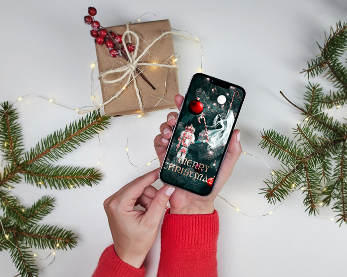 Christmas card mockup depicting a Christmas love story presented on a mobile screen, held in hands by a woman. Behind there are Christmas tree branches, a present and Christmas tree lights