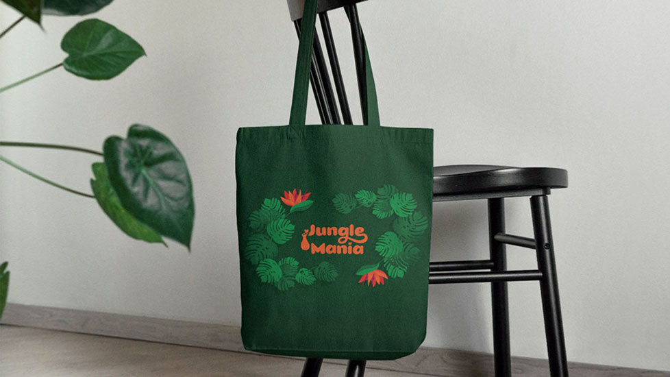 On a chair there is hanged a tote bag depicting the a tropical illustrations with Monstera Deliciosa leaves and Bird of paradise flowers and the logotype of Jungle Mania. Next to it, there are visible some leaves.