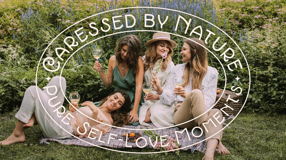 Caressed by Nature submark logo design depicted on top of the image of a group of young women smiling and holding wine glasses at a picnic in nature